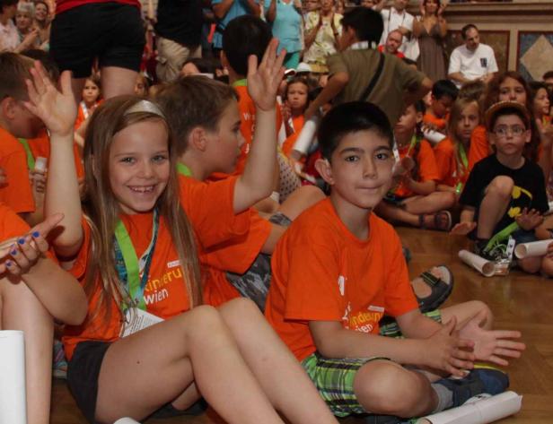 Eine Gruppe Kinder sitzt auf dem Boden und winkt in die Kamera.