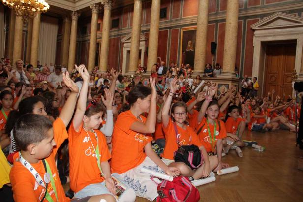 Eine Gruppe Kinder in orangefarbenen T-Shirts sitzt in einem Saal und hebt die Hände.