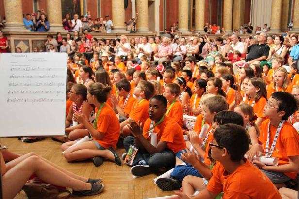Eine Gruppe Kinder in orangefarbenen T-Shirts sitzt auf dem Boden und klatscht Beifall.