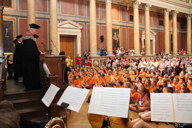 Eine Gruppe Kinder in orangefarbenen T-Shirts sitzt vor einer Bühne mit Rednern.