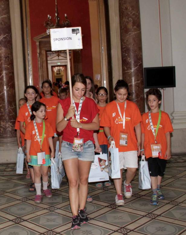 Eine Gruppe Kinder in orangefarbenen T-Shirts geht mit einer jungen Frau, die ein Schild mit der Aufschrift „Sponsion“ trägt.