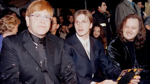 Elton John, David Beckham und ein dritter Mann sitzen nebeneinander bei einer Veranstaltung.