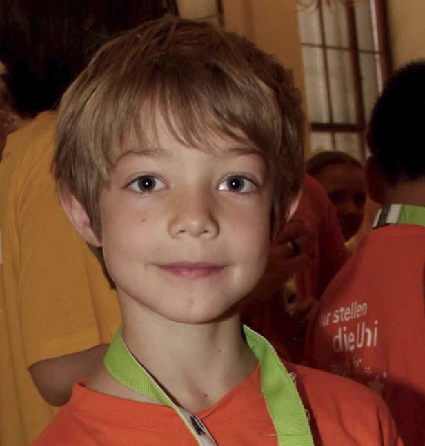 Ein junger Junge mit blonden Haaren und einem orangenen T-Shirt lächelt.