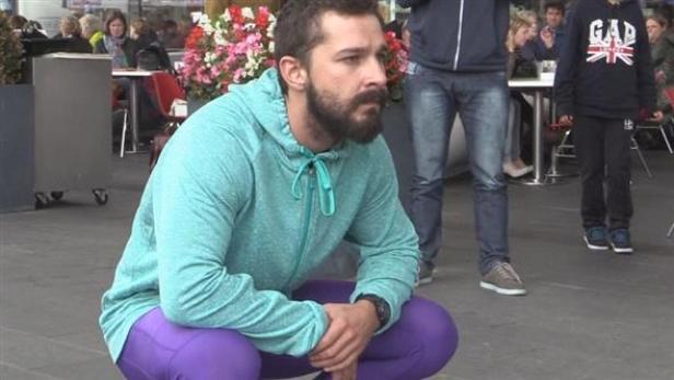 Shia LaBeouf hockt in einem türkisfarbenen Hoodie und lila Leggings auf einem Platz.