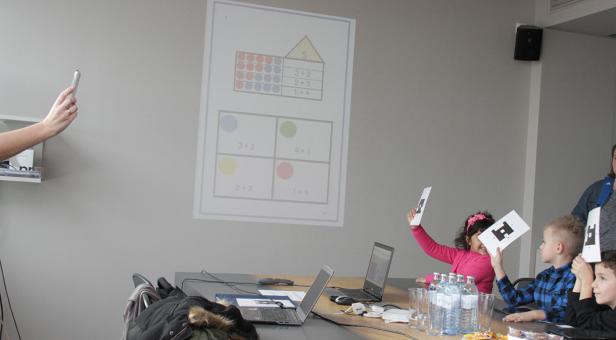 Kinder sitzen an einem Tisch und lernen mit einer Projektion an der Wand Rechnen.