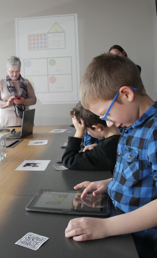 Ein Junge mit Brille tippt auf einem Tablet, im Hintergrund eine Tafel mit Rechenaufgaben.