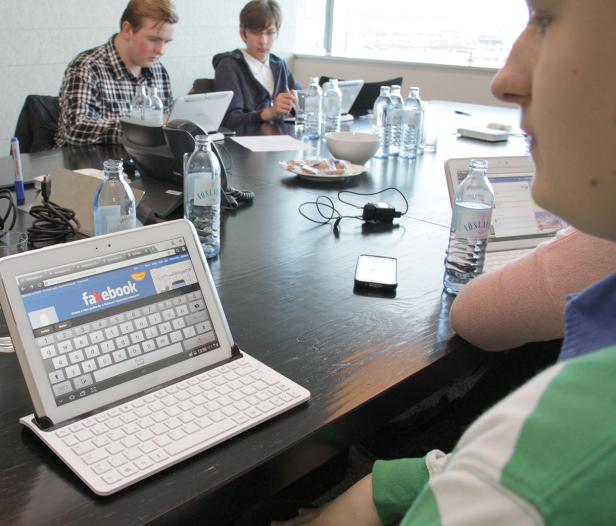 Mehrere Personen sitzen an einem Tisch mit Laptops und Tablets, auf einem Tablet ist „Facebook“ zu sehen.