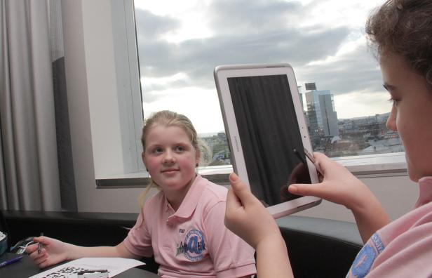 Ein Mädchen hält ein Tablet vor ein anderes Mädchen in einem Raum mit Blick auf eine Stadt.