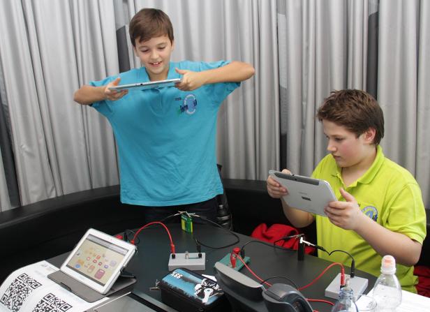 Zwei Jungen experimentieren mit Tablets und elektronischen Bauteilen auf einem Tisch.
