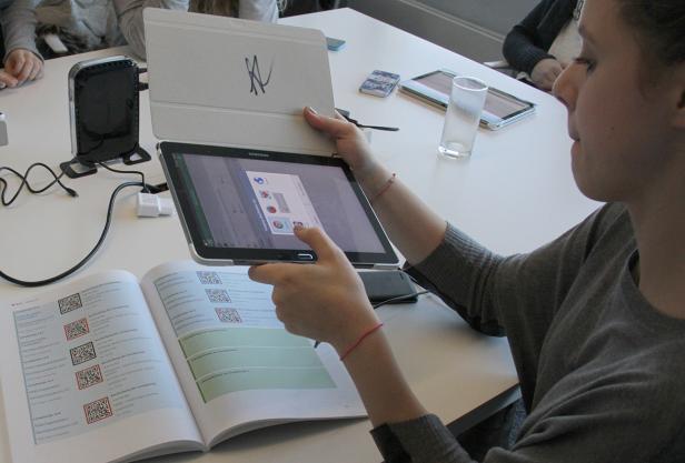 Eine Person hält ein Tablet über ein aufgeschlagenes Buch mit QR-Codes.