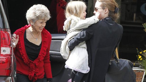 Eine Frau mit Sonnenbrille trägt ein kleines Mädchen, während eine ältere Dame daneben steht.