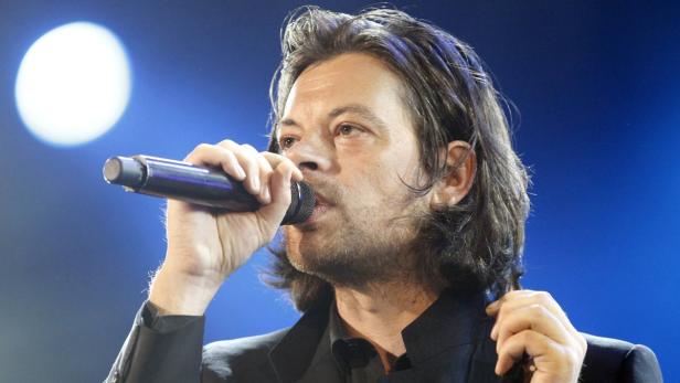 Ein Mann mit dunklen Haaren singt in ein Mikrofon vor blauem Hintergrund.