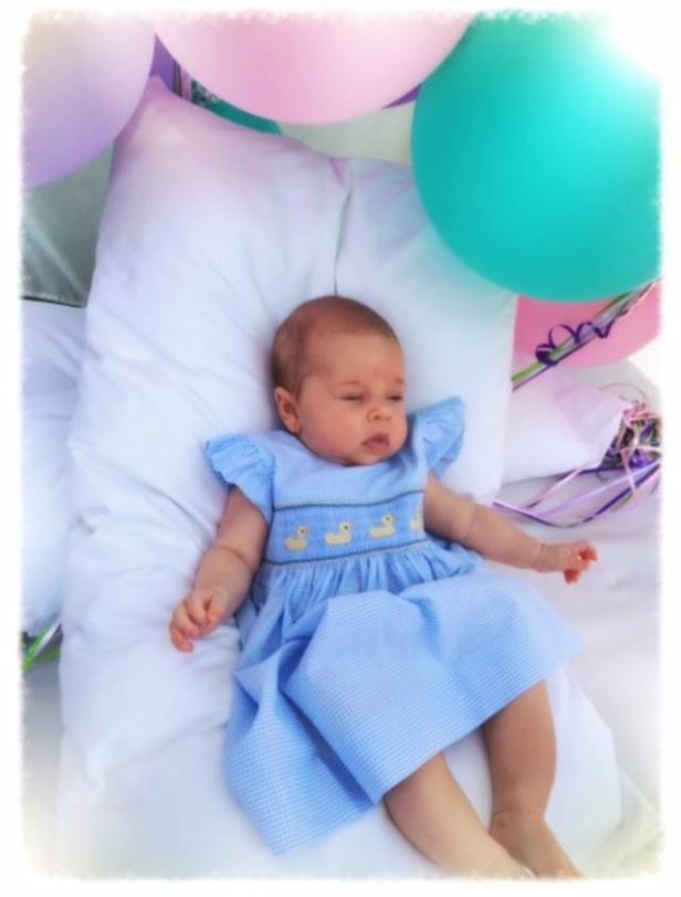 Ein Baby in einem blauen Kleid mit Entenmotiven liegt auf einem Kissen, umgeben von Ballons.
