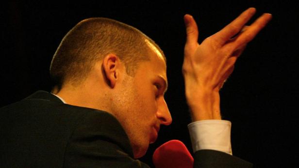 Ein Mann im Anzug gestikuliert mit der Hand vor einem dunklen Hintergrund.