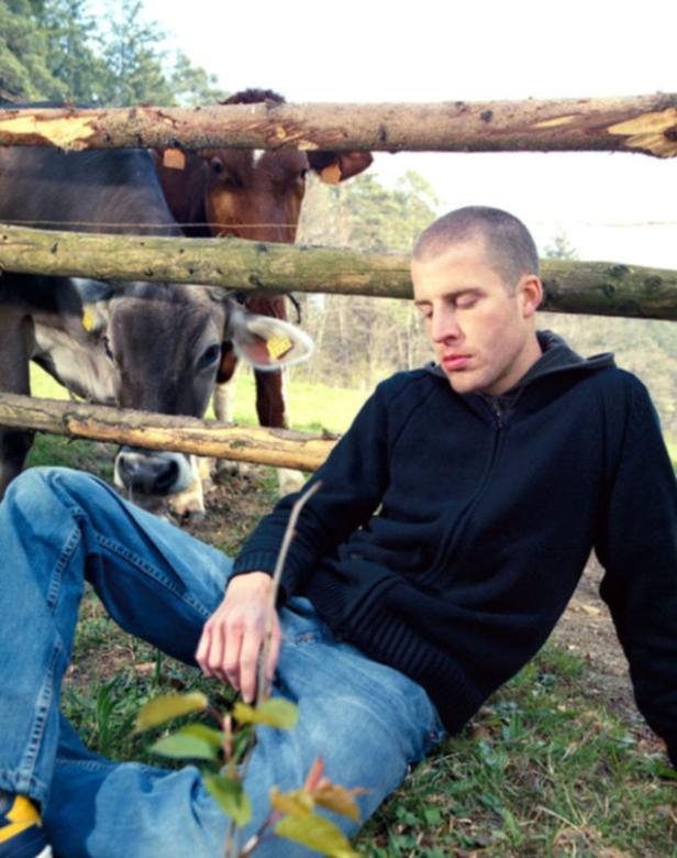Ein Mann sitzt auf einer Wiese vor einem Holzzaun, hinter dem Kühe stehen.