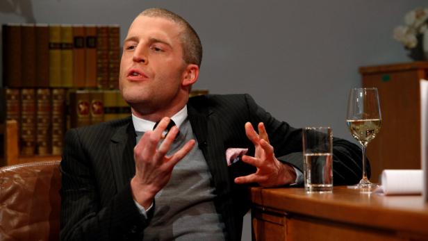 Ein Mann im Anzug gestikuliert an einem Tisch mit Getränken.