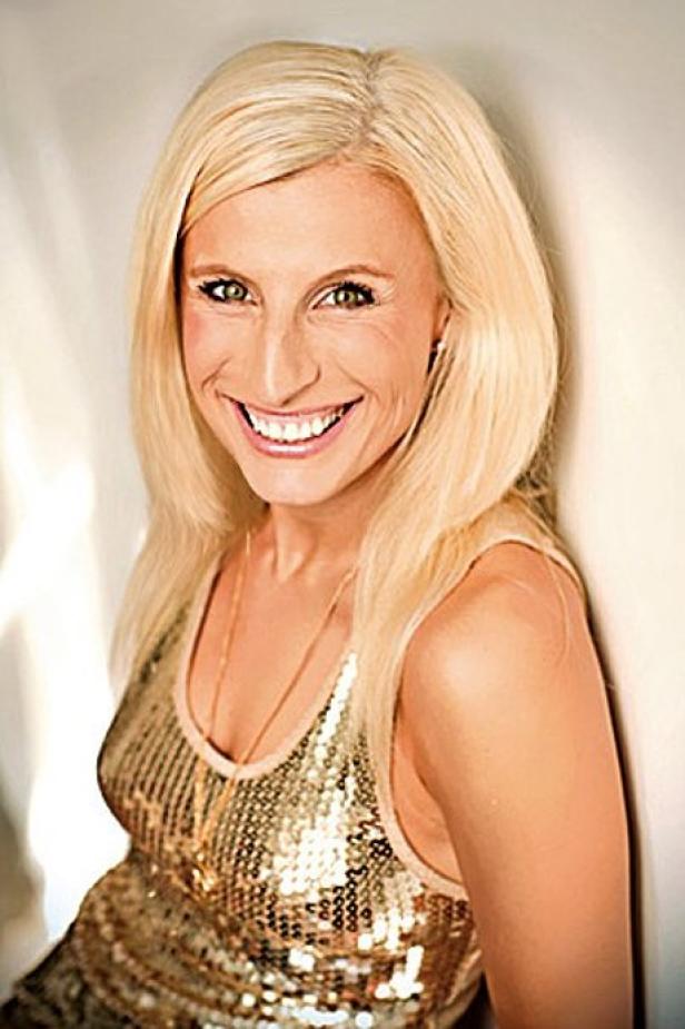 Eine lächelnde Frau mit blonden Haaren und einem goldenen Paillettenoberteil lehnt an einer Wand.