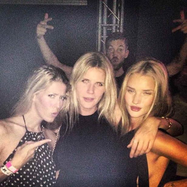 Drei blonde Frauen posieren für ein Foto, während zwei Männer im Hintergrund Grimassen schneiden.