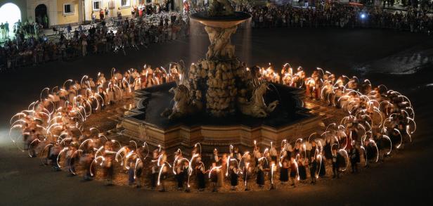 Eine Gruppe von Menschen in Tracht umrundet einen Brunnen mit Fackeln in der Nacht.