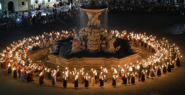Menschen in Tracht umringen den Brunnen auf dem Residenzplatz in Salzburg mit Kerzen.