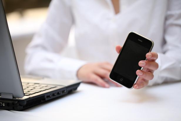 Eine Person hält ein Smartphone in der Hand, während ein Laptop auf dem Tisch steht.