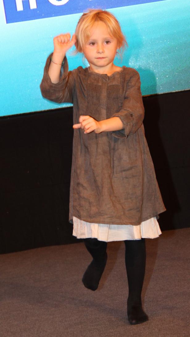 Ein kleines Mädchen in einem grauen Kleid tanzt auf einer Bühne.