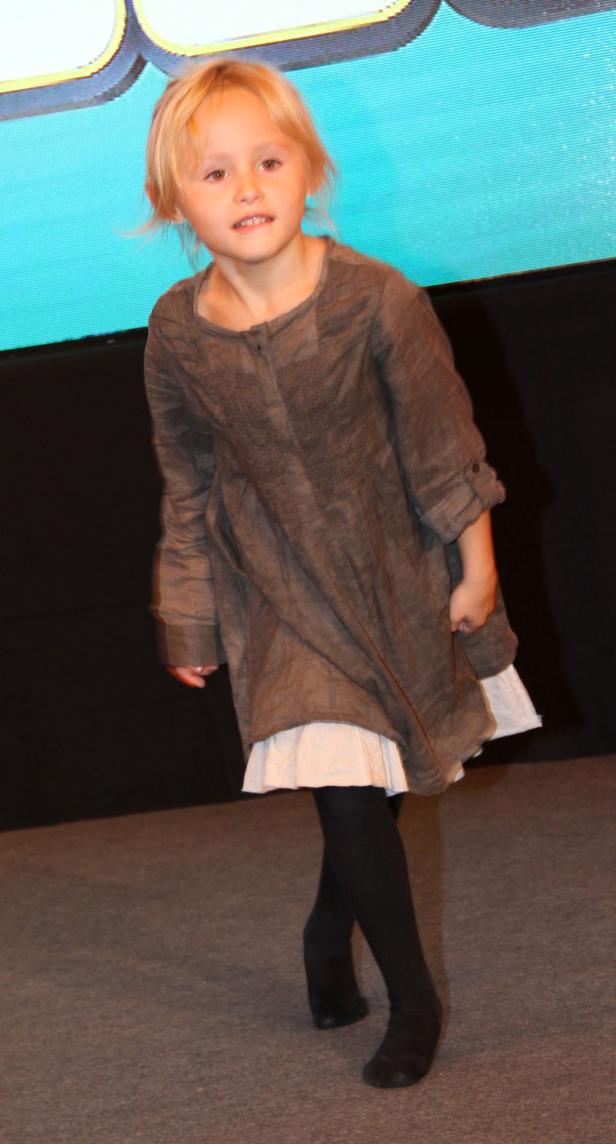 Ein blondes Mädchen in einem braunen Kleid und schwarzen Strumpfhosen steht auf einer Bühne.