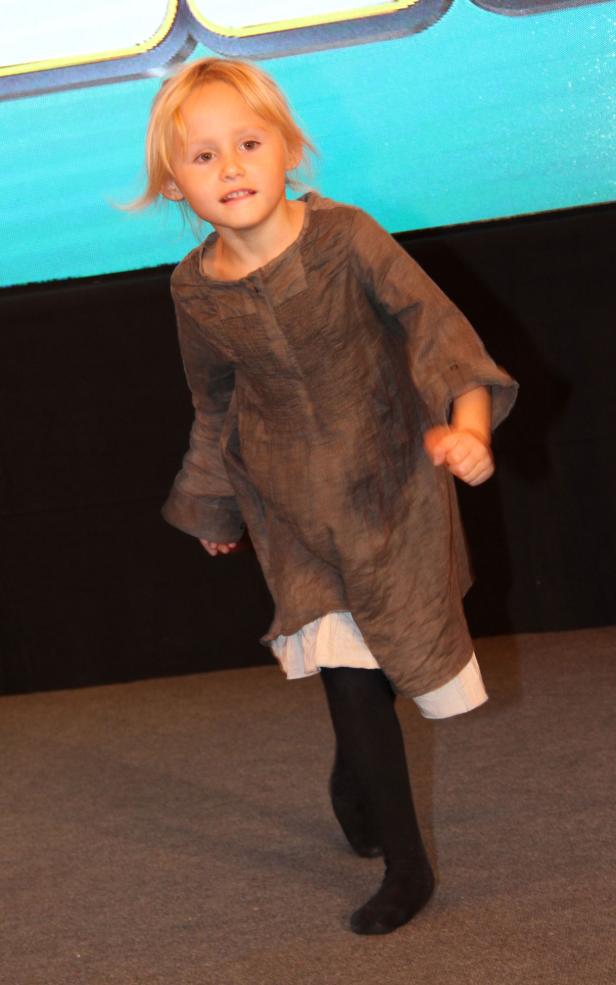 Ein kleines Mädchen in einem braunen Kleid steht auf einer Bühne.