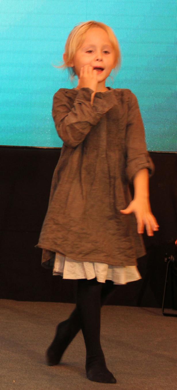 Ein blondes Mädchen in einem braunen Kleid steht auf einer Bühne.