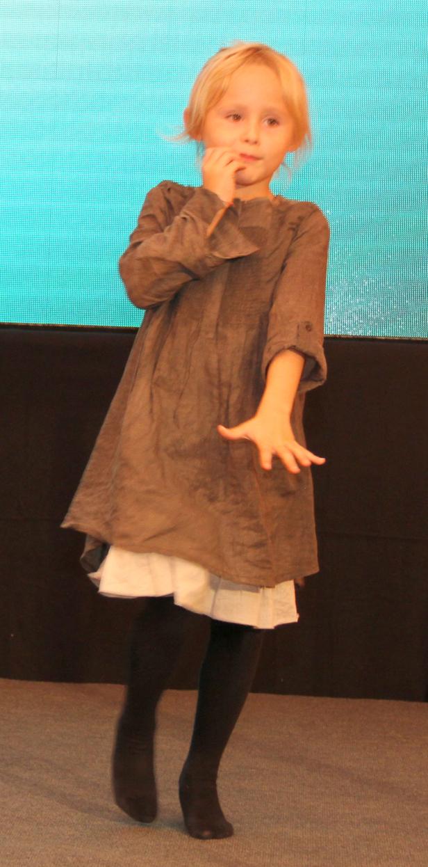 Ein blondes Mädchen in einem braunen Kleid tanzt vor einer blauen Wand.