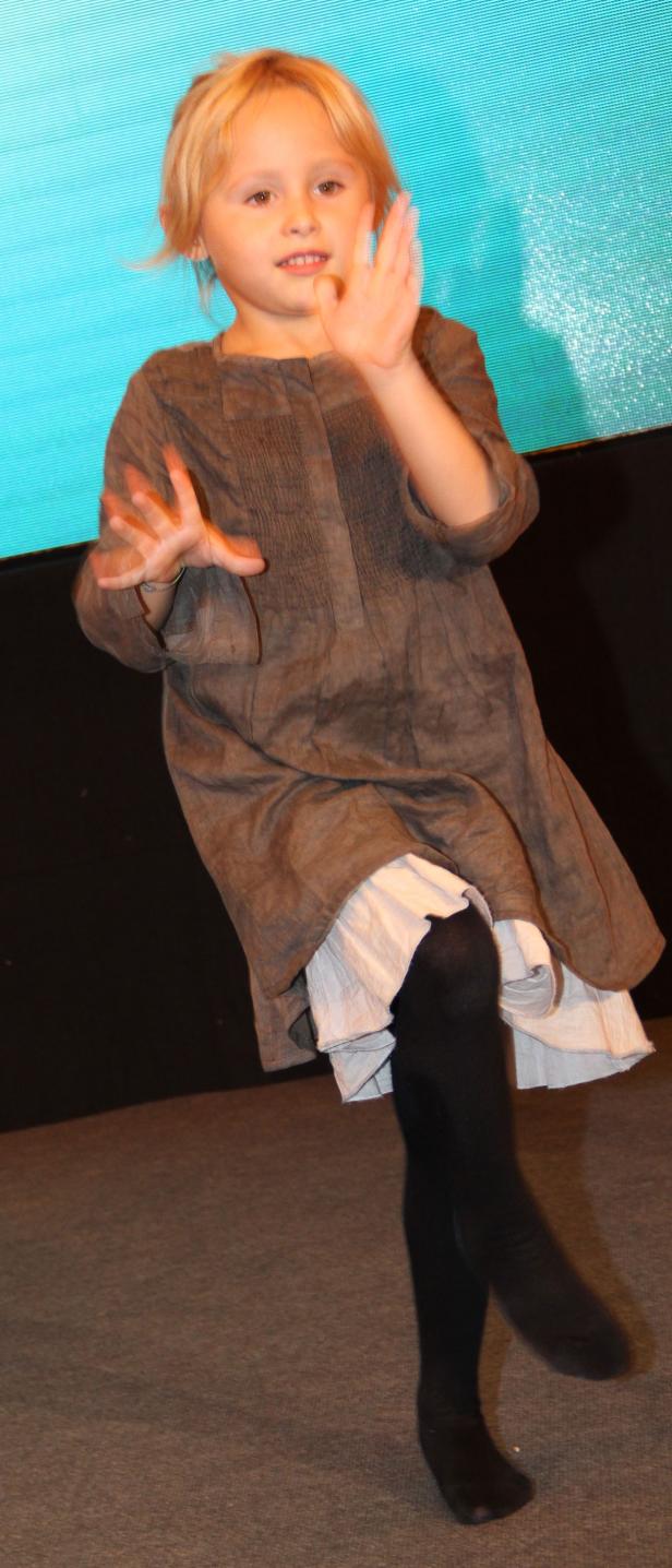 Ein kleines Mädchen in einem braunen Kleid tanzt vor einer blauen Wand.