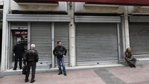 Vor einer geschlossenen Bank stehen ein Mann am Geldautomaten, ein Wartender und eine sitzende, obdachlose Person.