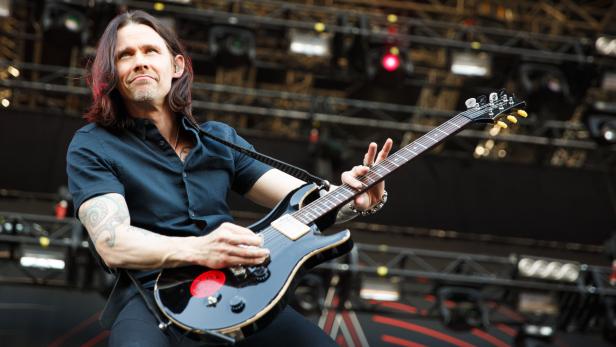 Ein Mann mit Tätowierungen spielt Gitarre auf einer Bühne.