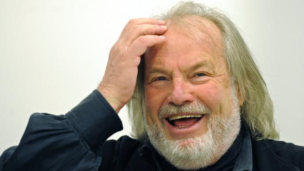 Ein lachender Mann mit grauem Haar und Bart berührt seine Stirn.