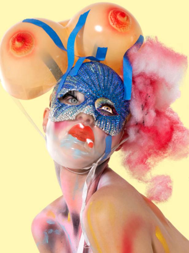 Eine Person mit einer glitzernden Maske und Ballons in Form von Brüsten vor einem gelben Hintergrund.