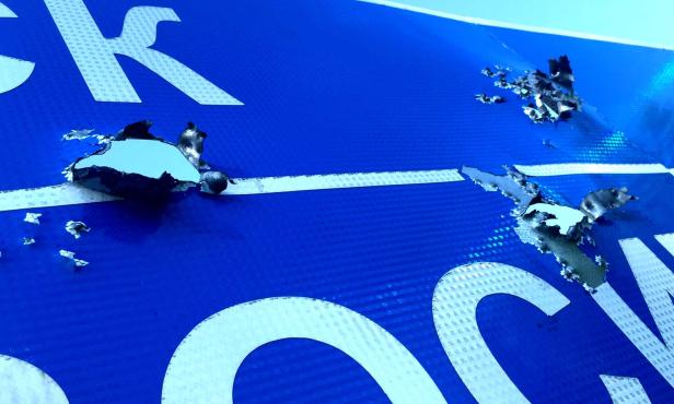 Ein beschädigtes blaues Schild mit weißen Buchstaben und abgerissenen Stellen.