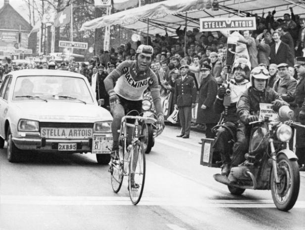 Eddy Merckx fährt ein Radrennen, begleitet von Zuschauern und Begleitfahrzeugen.
