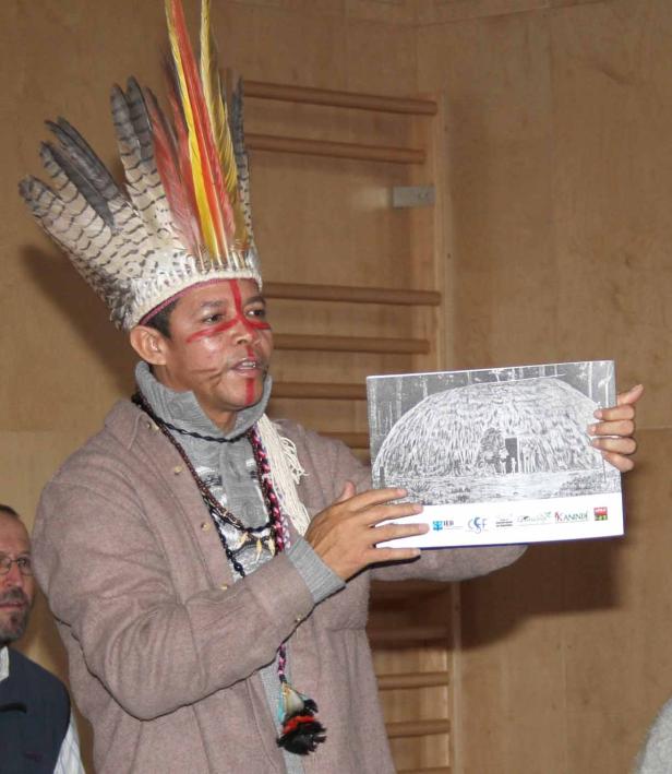 Ein Mann mit Kopfschmuck und Gesichtsbemalung präsentiert ein Foto eines traditionellen Hauses.