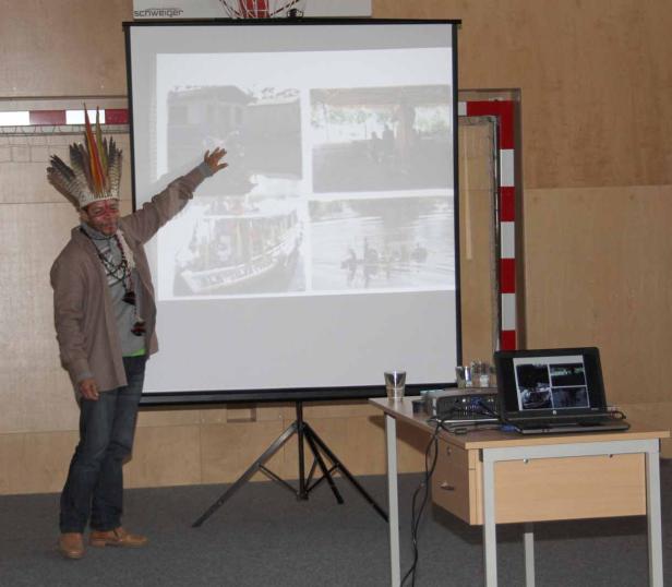 Ein Mann mit Kopfschmuck präsentiert Bilder von Booten und Flüssen.