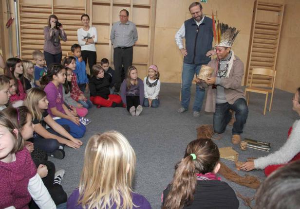 Ein Mann mit Kopfschmuck zeigt einer Gruppe von Kindern einen Korb.