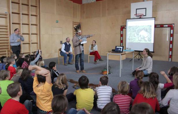 Ein Mann mit Kopfschmuck hält einen Vortrag vor einer Gruppe von Kindern.