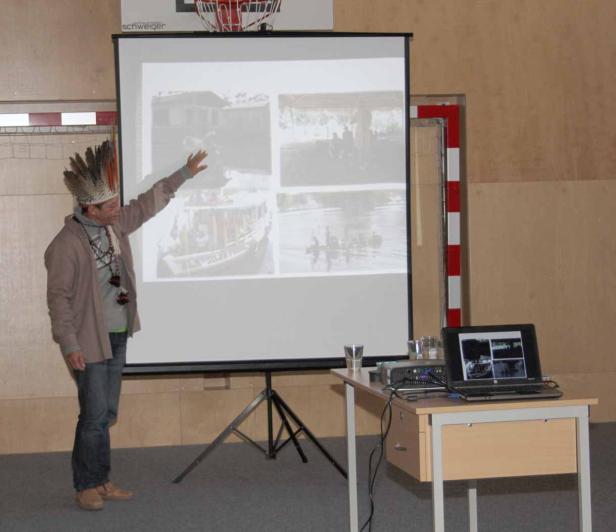 Ein Mann mit Federschmuck präsentiert Fotos von einer Reise auf einer Leinwand.