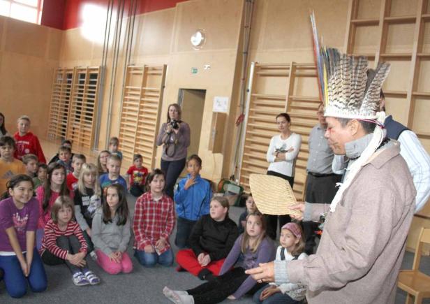 Ein Mann mit Federschmuck spricht vor einer Gruppe von Schulkindern in einer Turnhalle.