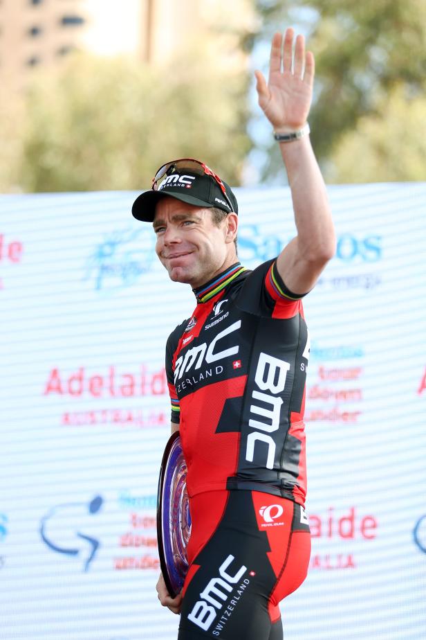 Ein Radrennfahrer in roter und schwarzer BMC-Teamkleidung winkt mit einem Pokal in der Hand.