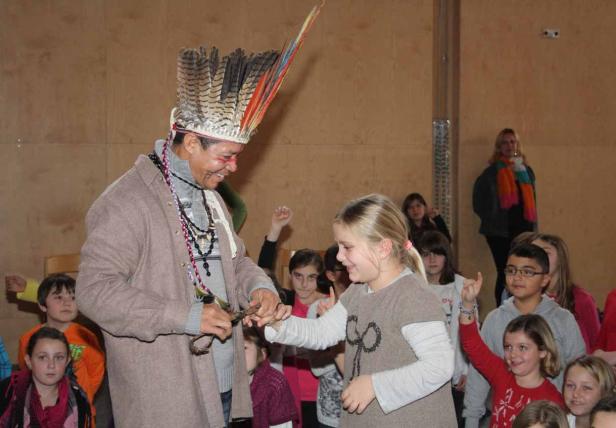 Ein Mann mit Federschmuck überreicht einem Mädchen ein Geschenk vor einer Gruppe von Kindern.