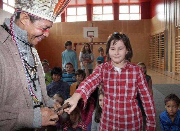 Ein Mann mit Kopfschmuck reicht einem Mädchen in einem roten Hemd einen Gegenstand.