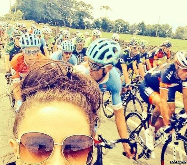 Eine Frau macht ein Selfie vor einer Gruppe von Radfahrern bei einem Rennen.