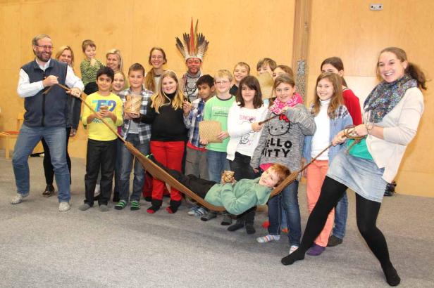 Eine Gruppe von Kindern und Erwachsenen hält eine Hängematte, in der ein Junge liegt.