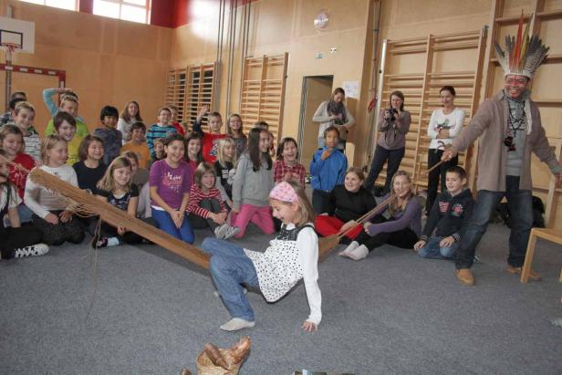 Eine Schulklasse beobachtet, wie ein Mädchen unter einer Stange hindurchtanzt, angeleitet von einem Mann mit Federschmuck.