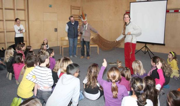 Eine Gruppe Kinder sitzt vor zwei Männern und einer Frau, die eine Art Netz präsentieren.